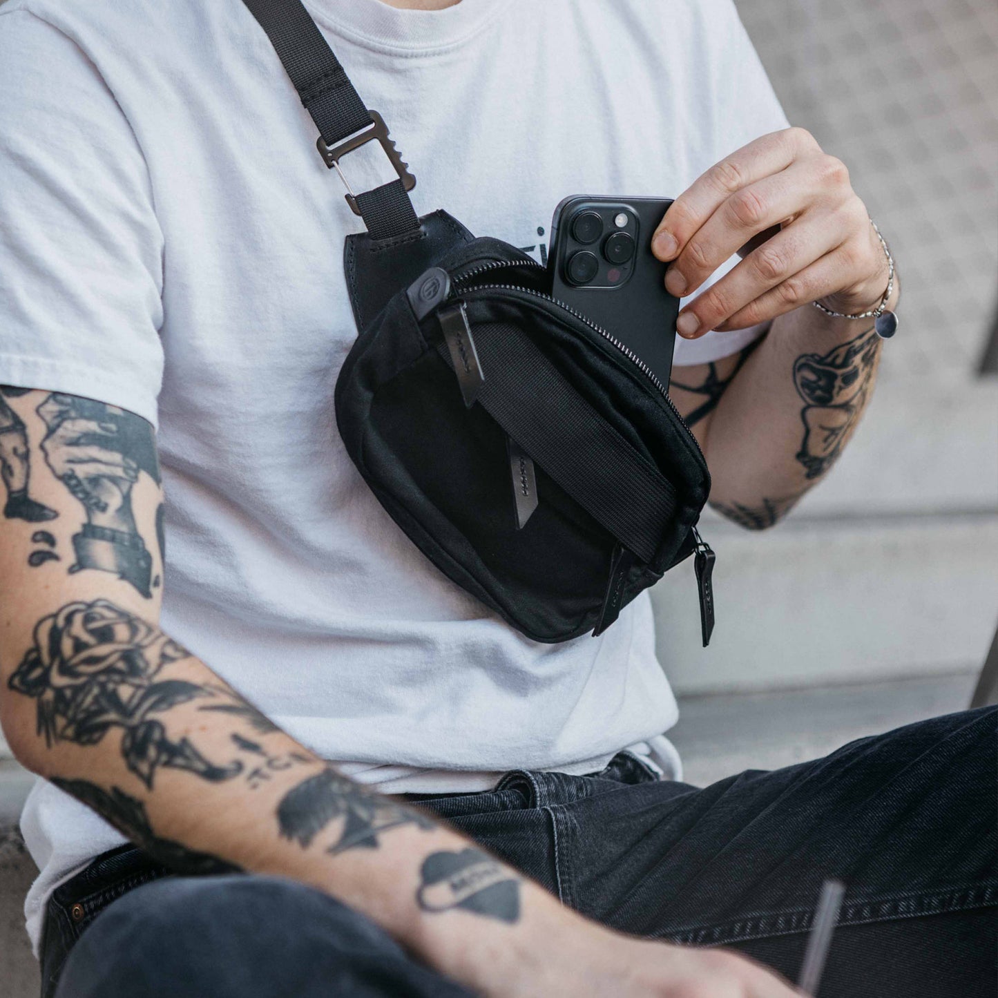 Slate Grey Waxed Canvas Canyon Crossbody Mini, worn across the chest by a person putting a black smartphone into its zippered compartment.