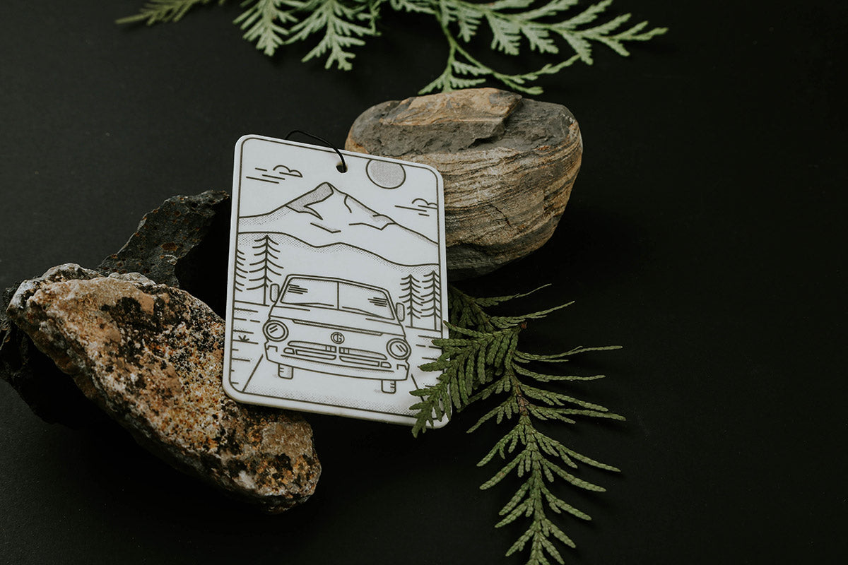Black and white illustrated air freshener by Tanner Goods featuring a camper van in mountains and pines, styled with rocks and foliage.