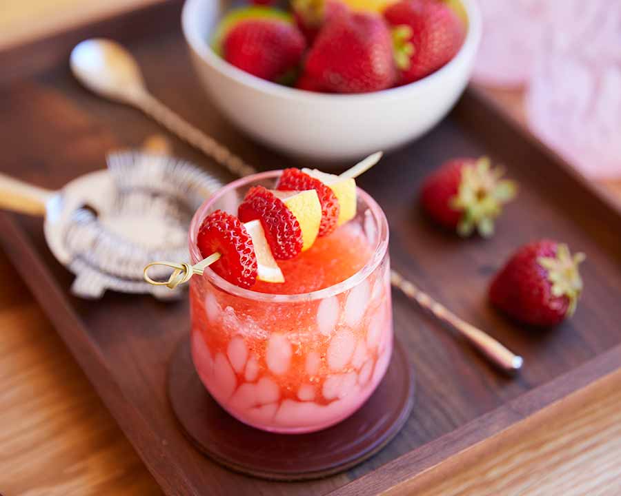 Vibrant pink slushie garnished with strawberries and pineapple, served on a wooden tray with fresh berries and a gold spoon.