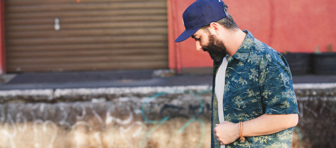 Bearded man in blue cap and patterned shirt walks thoughtfully past graffiti-covered wall, embodying urban calm.