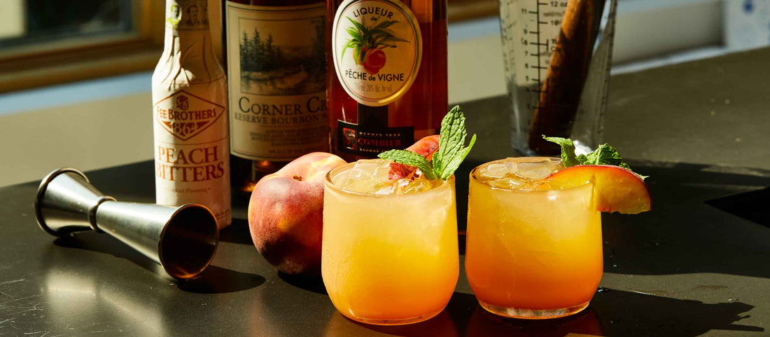 Two vibrant peach cocktails garnished with mint and peach slices, surrounded by bottles of bourbon, peach liqueur, and bitters on a sunlit bar.