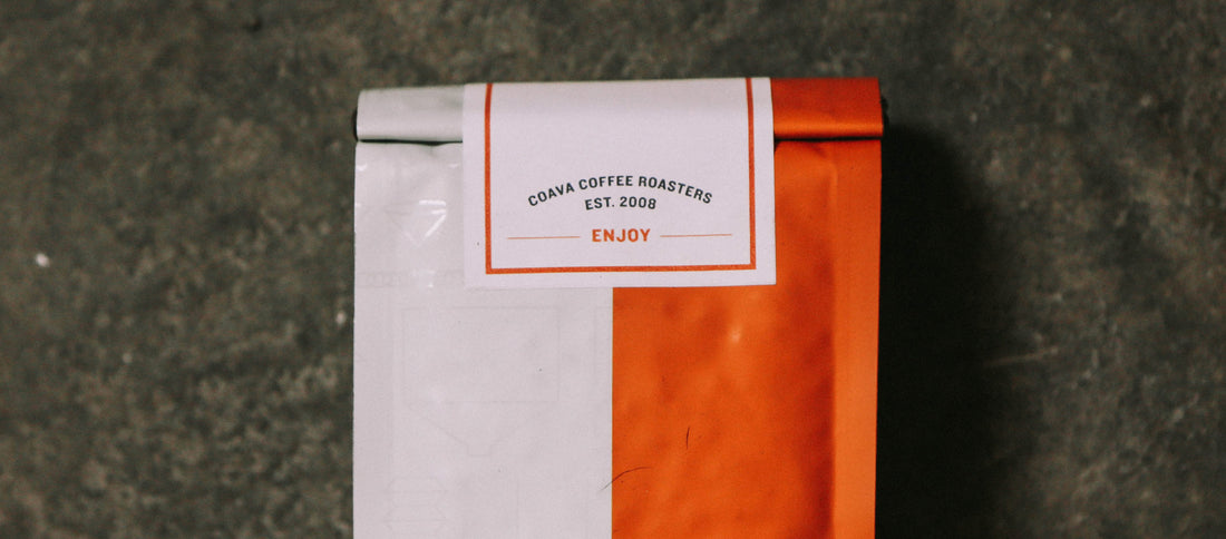 White and orange Coava Coffee Roasters bag with "Enjoy" label, resting on dark textured surface.