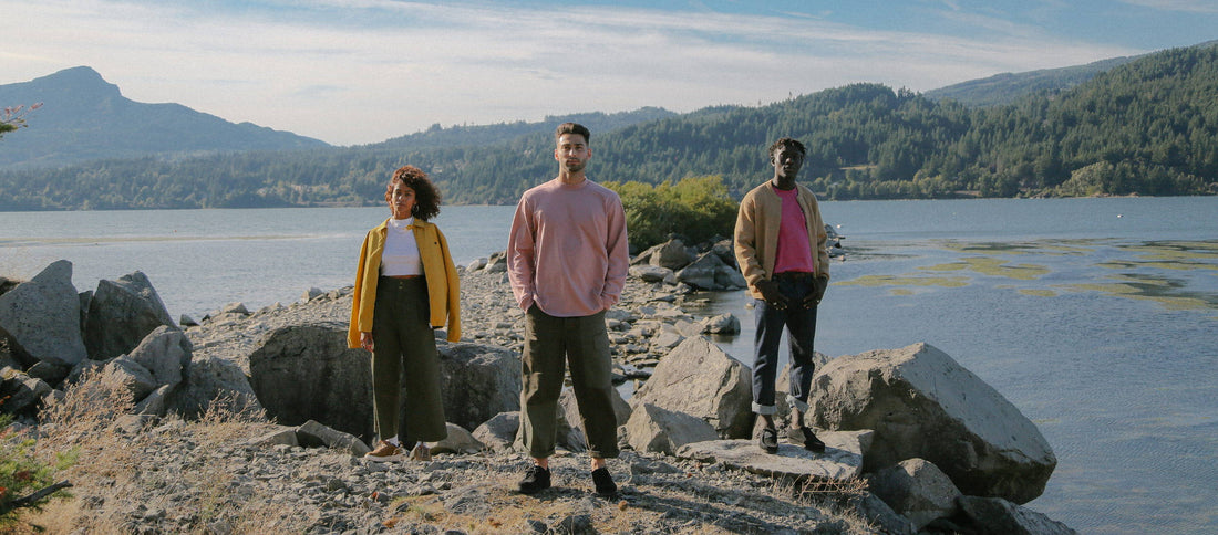 Three models in Fall 19 outfits stand confidently on rocky shores by a calm lake, surrounded by forested hills under soft sunlight.