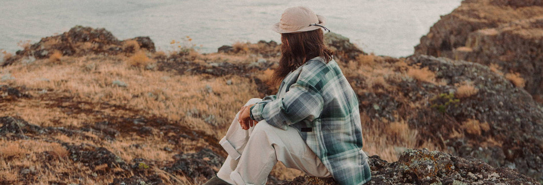 Person in plaid shirt and hat sits contemplatively on rocky cliff overlooking calm ocean, evoking reflection and connection.