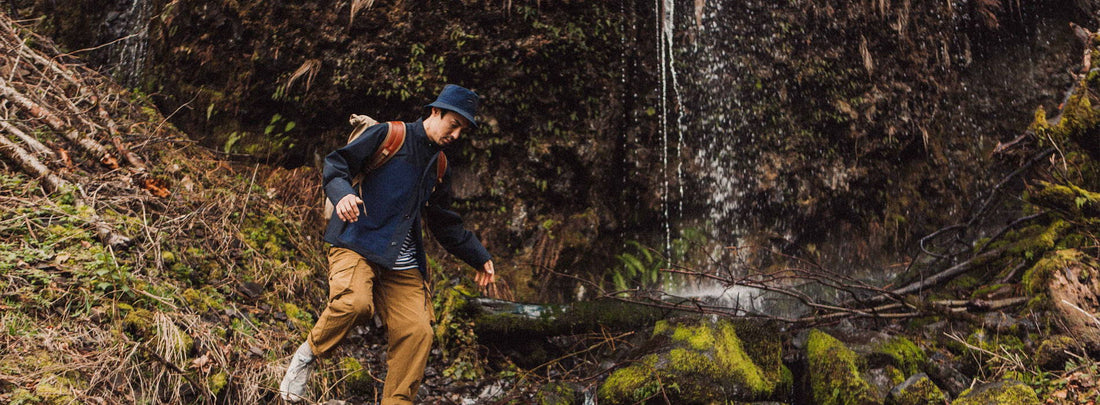 Man in Nanamica gear hikes through mossy forest, waterfall behind, embodying outdoor exploration spirit.