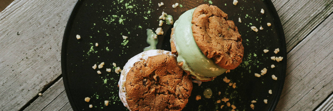 Two ice cream sandwiches with chocolate chip cookies and green and white ice cream, served on a black plate with crumbles, on a rustic wooden table.