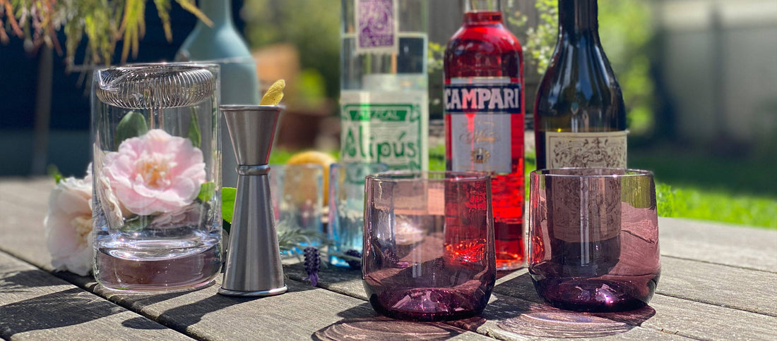 Sunlit backyard bar setup with mezcal, Campari, and purple glasses, fresh flowers, and cocktail tools for a relaxed at-home Negroni.
