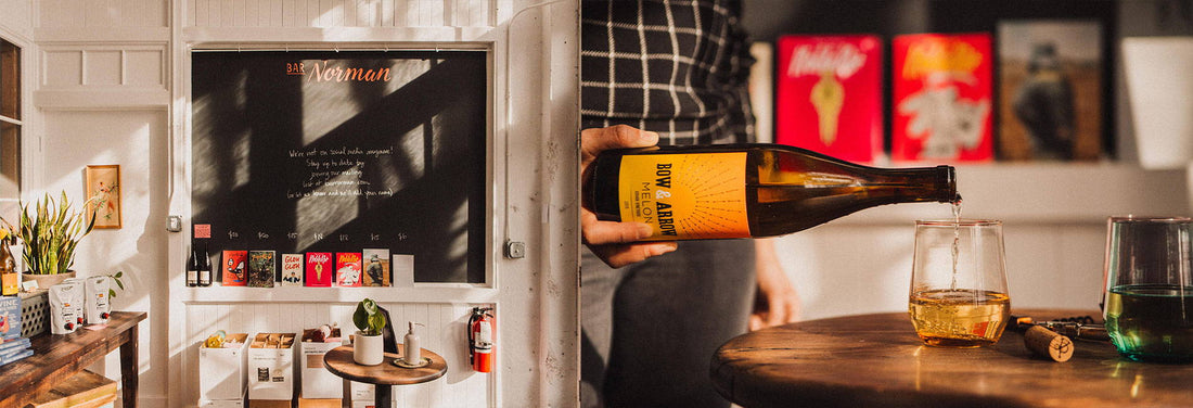 Person pouring golden wine into glass at Bar Norman, cozy shop with chalkboard sign and colorful art.