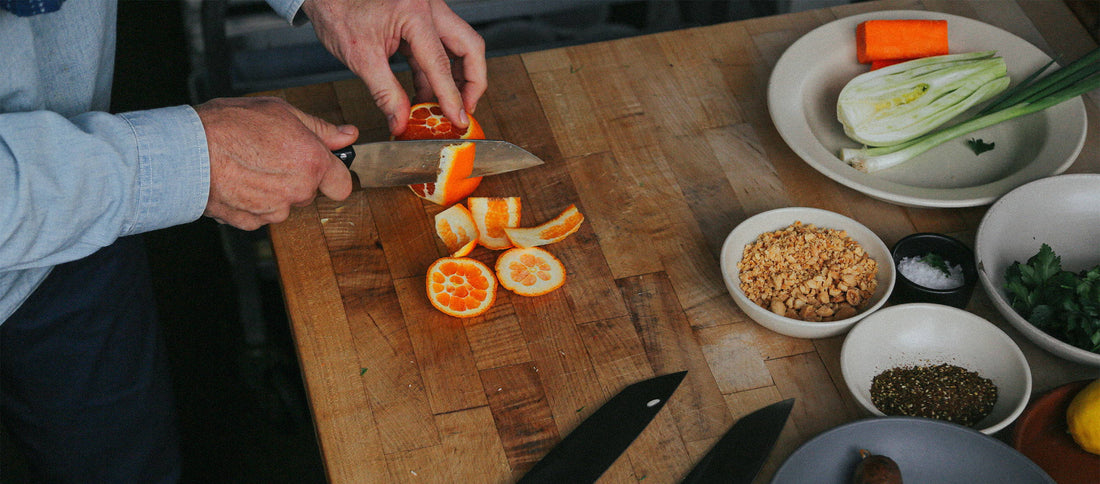 Chef's hands slicing orange on wooden board with fresh ingredients nearby, showcasing culinary precision and vibrant flavors.
