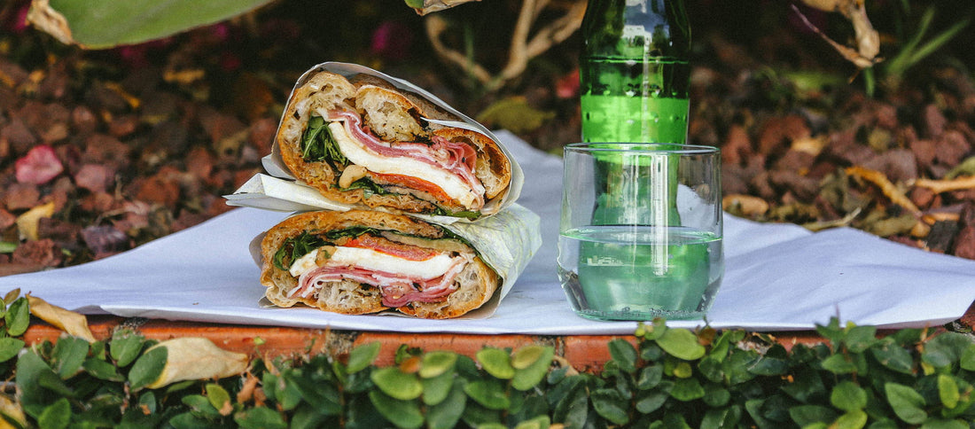 Freshly made Ggiata sandwich with prosciutto, mozzarella, and greens, served with sparkling water on a garden ledge.