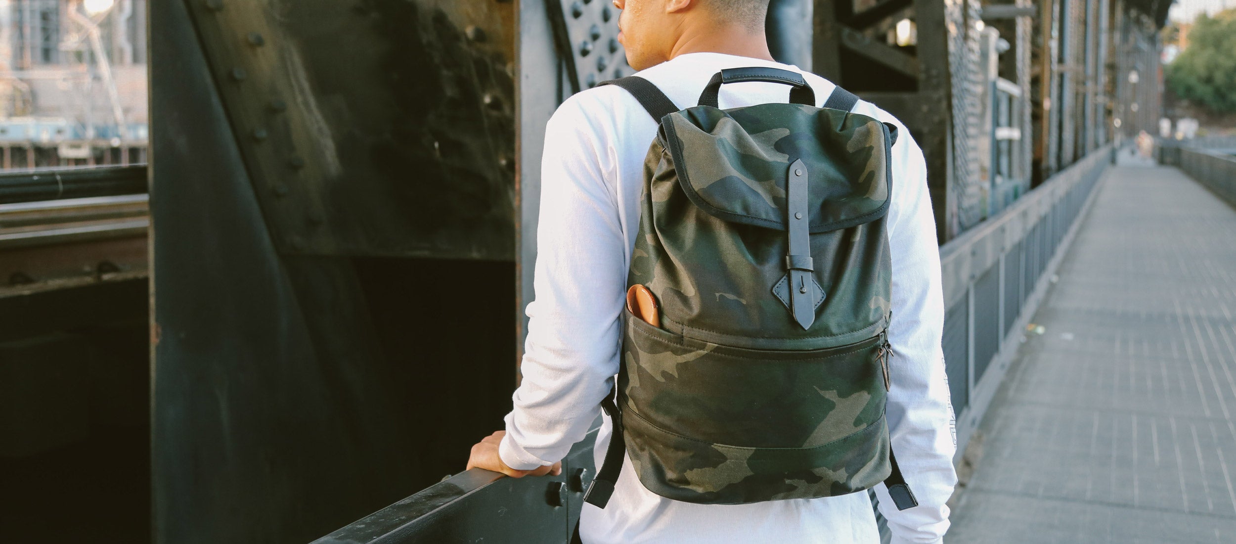 Man wearing M/81 Collection camouflage backpack, urban bridge walk, rugged style, military-inspired design.