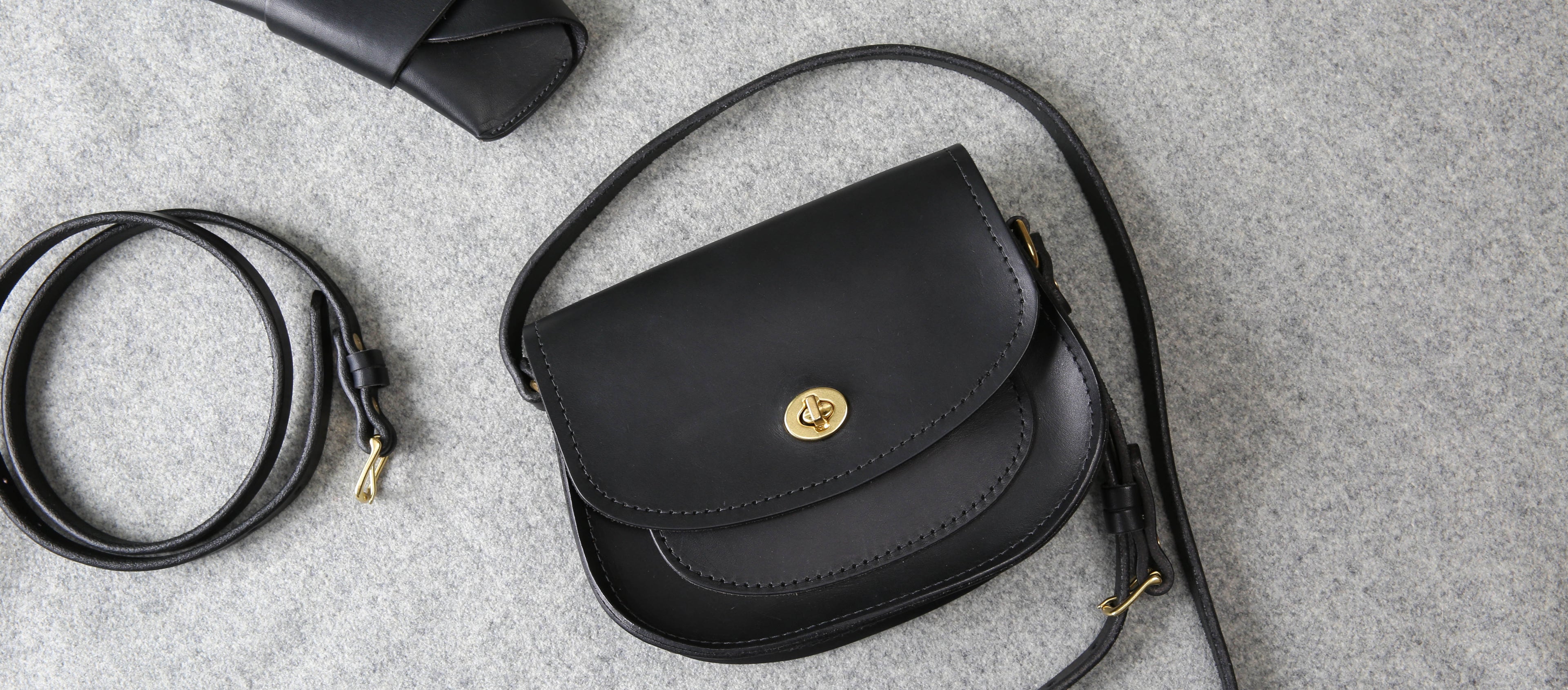 Elegant black leather crossbody bag with gold hardware, minimalist design, and soft gray backdrop.