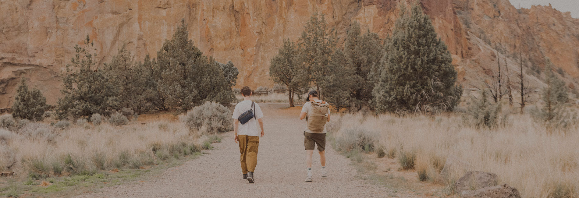 Two hikers in earth-toned outdoor gear walk a desert trail, backpacks and crossbody bags, surrounded by red rock and sagebrush.