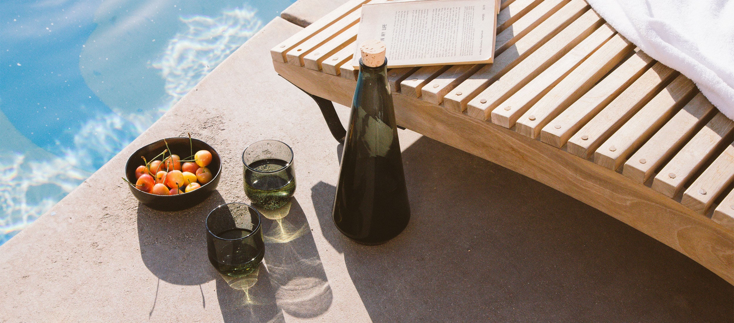 Elegant dark glass serving vessels, bowl, and stemless glasses by a poolside lounge, evoking relaxed, modern outdoor entertaining.