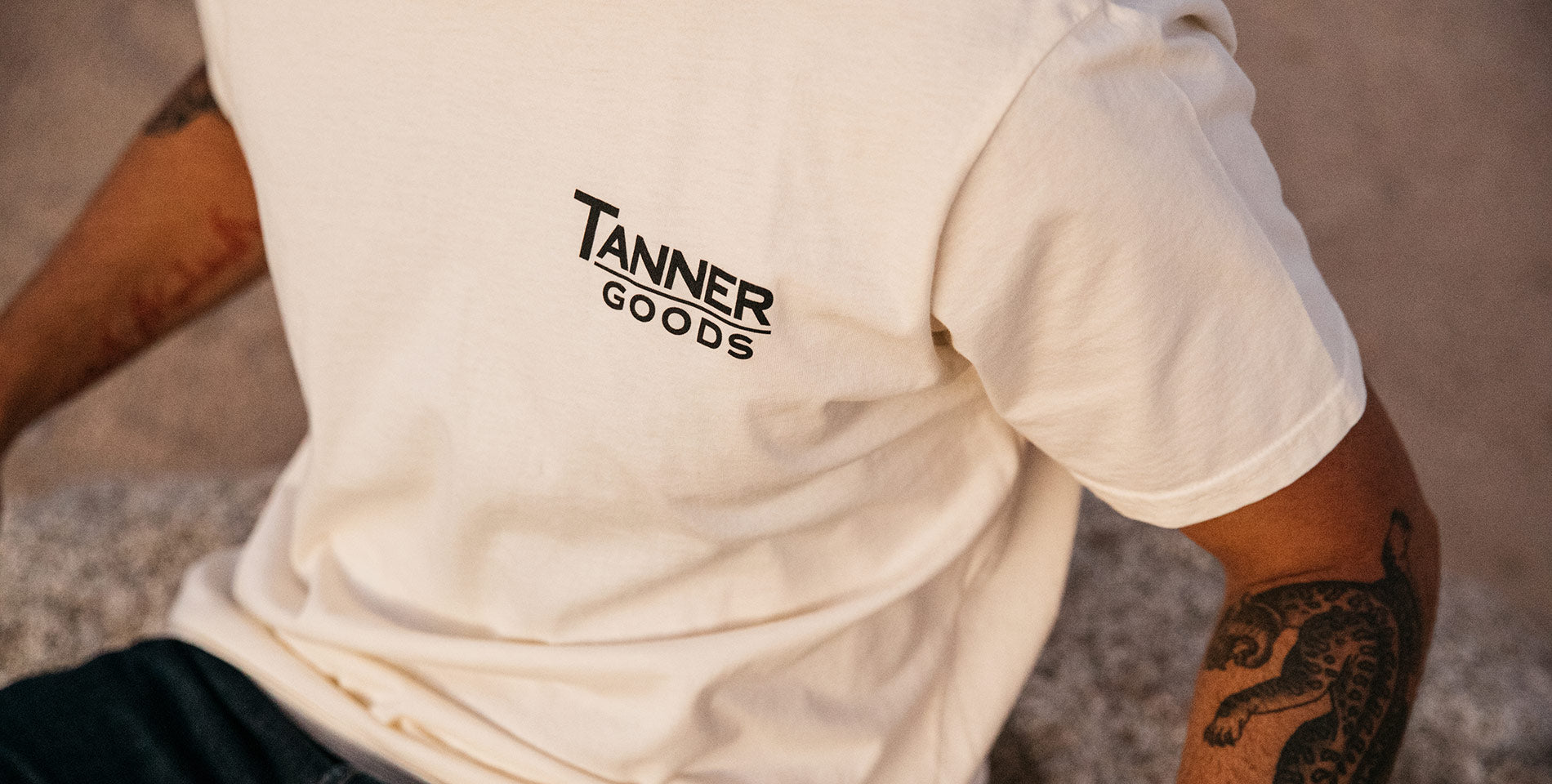 Cream T-shirt with bold "TANNER GOODS" logo, tattooed arms, minimalist urban style, soft texture, modern essentials.