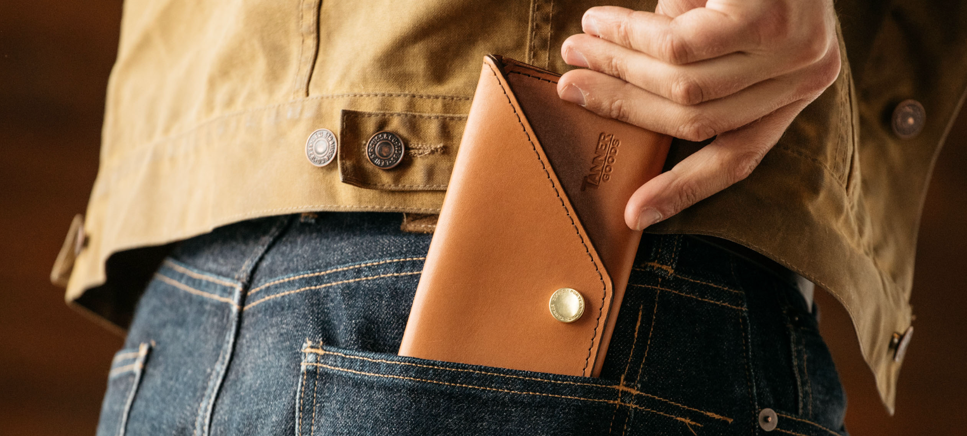 Tan leather wallet with brass snap, tucked into denim pocket, worn with rugged canvas jacket — durable, minimalist essentials for the modern craftsman.
