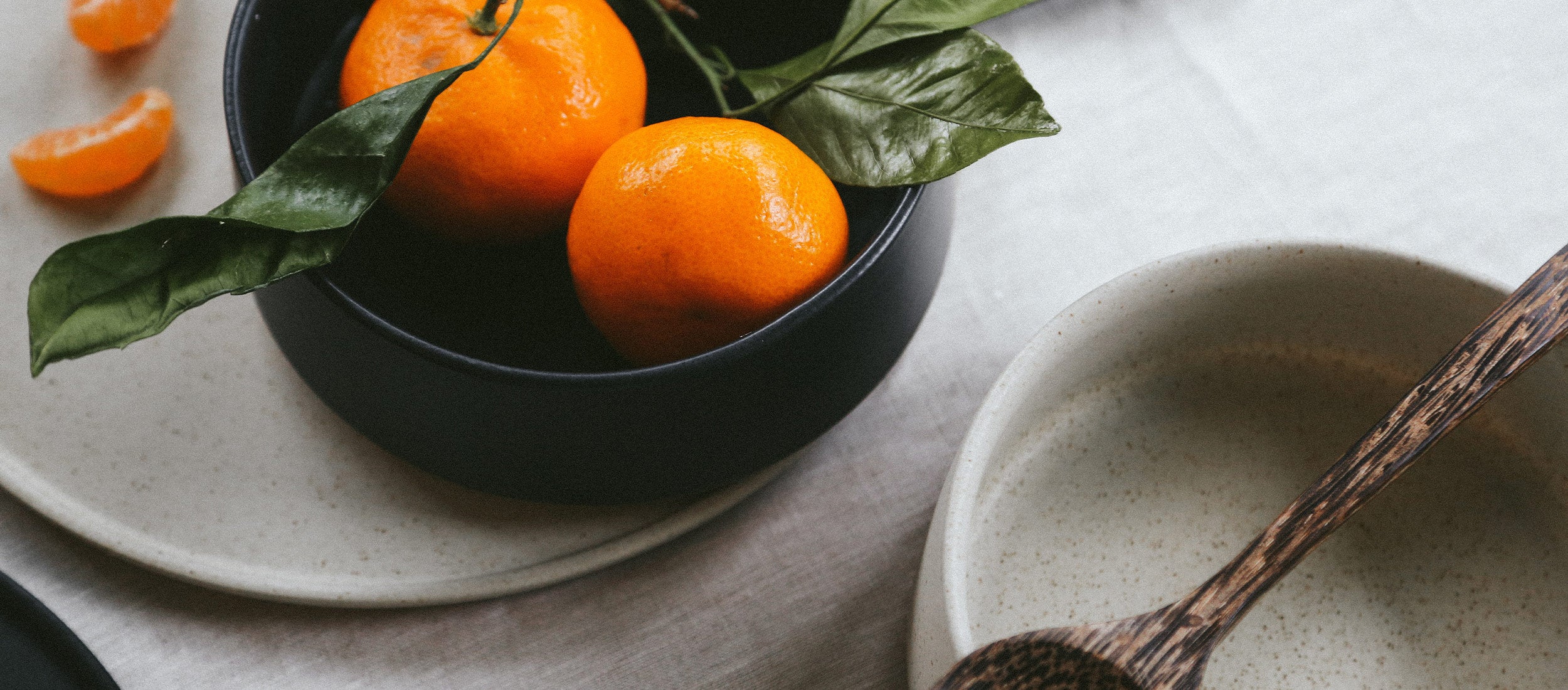 Minimalist ceramic bowls in black and speckled beige with fresh oranges and green leaves, evoking rustic elegance and natural simplicity.