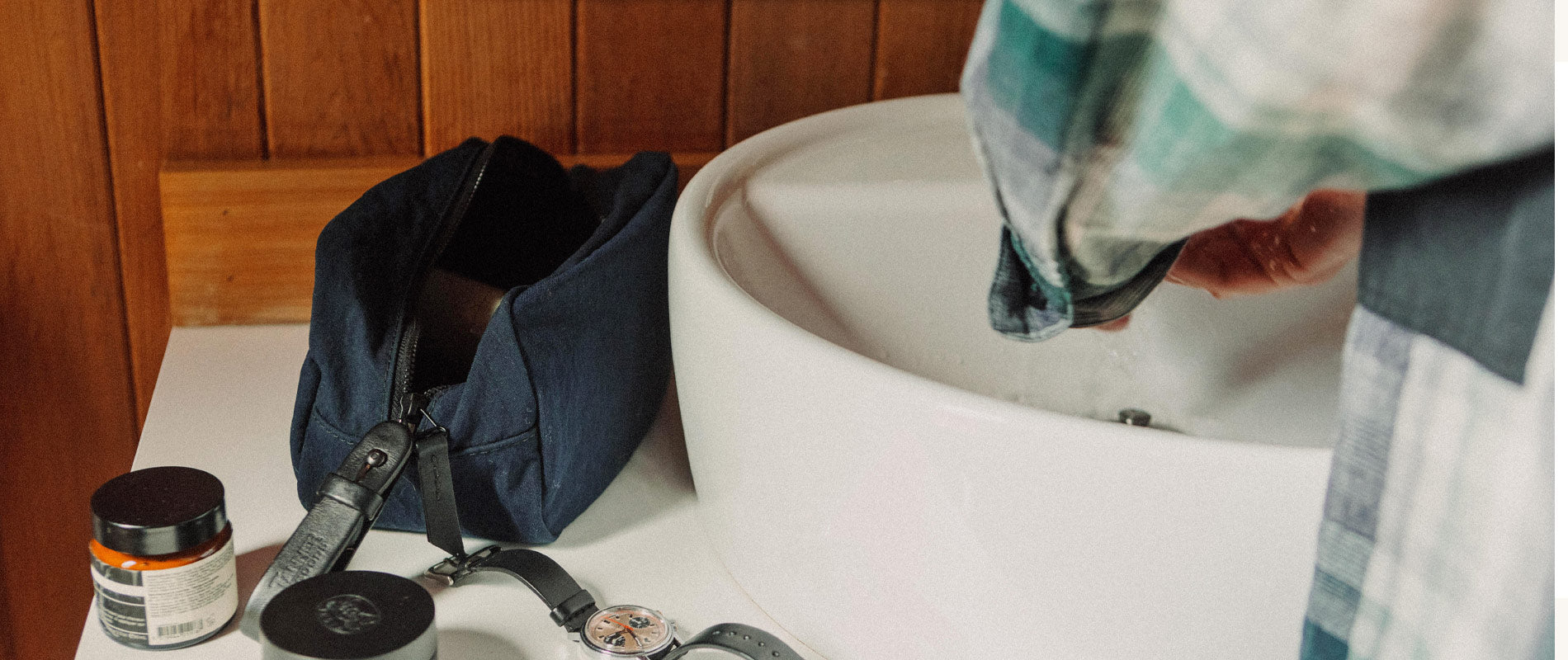 Apothecary collection: navy toiletry bag, amber jars, vintage watch, plaid robe—rustic wood, white sink, clean, minimalist self-care essentials.