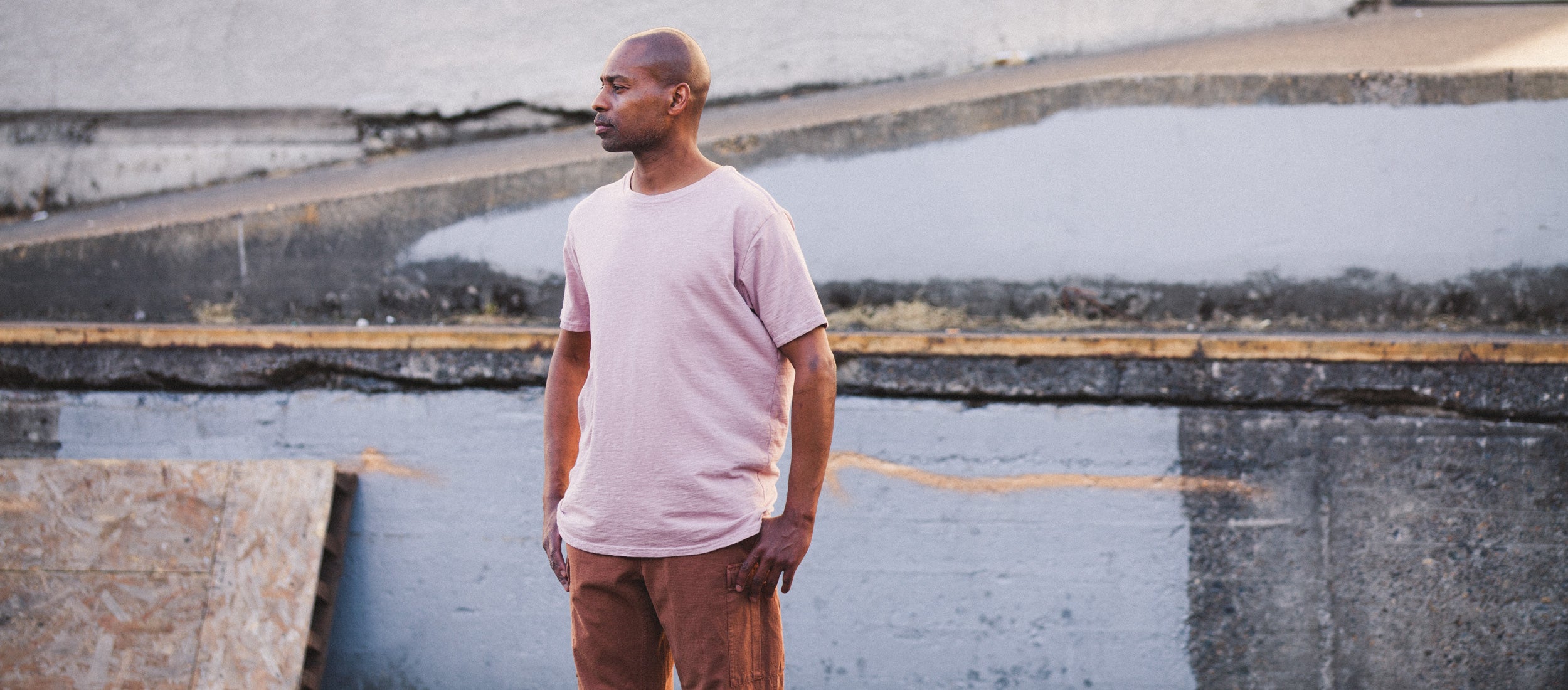 Man in soft pink tee and brown cargo pants stands by urban concrete, embodying Tanner Goods Essentials' minimalist, rugged style.