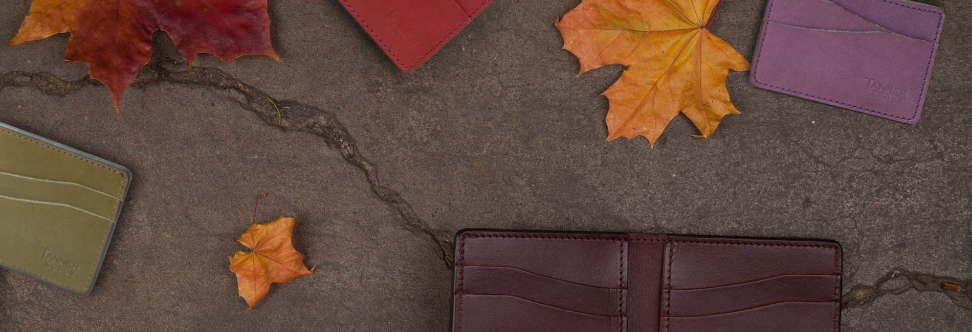 Rich leather wallets in burgundy, olive, red, and purple with autumn leaves on stone—2021 Holiday Limited Edition.
