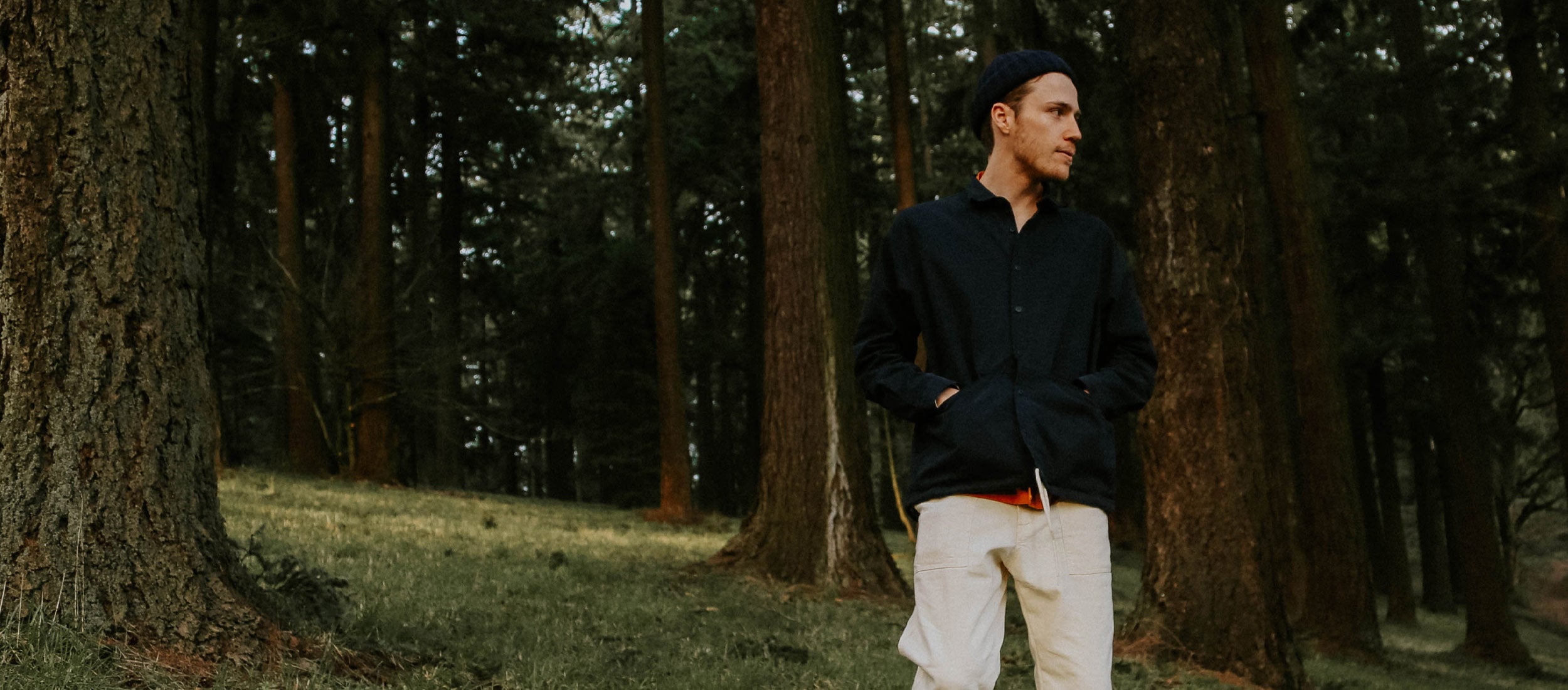 Man in navy shirt, cream pants, and beanie stands in forest, embodying UK Collection’s minimalist, nature-inspired style.