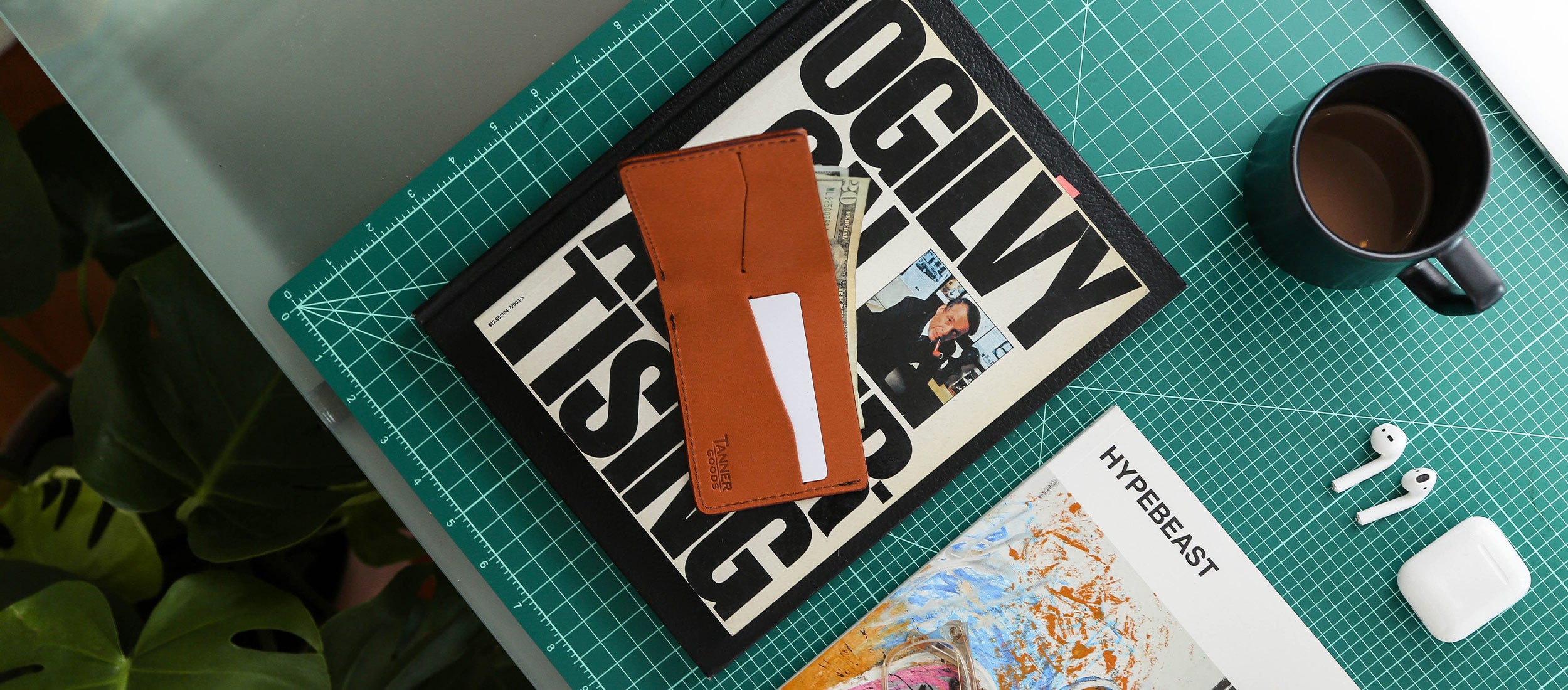 Slim tan leather wallet on green grid mat with magazines, coffee, and earbuds — minimalist urban lifestyle essentials.