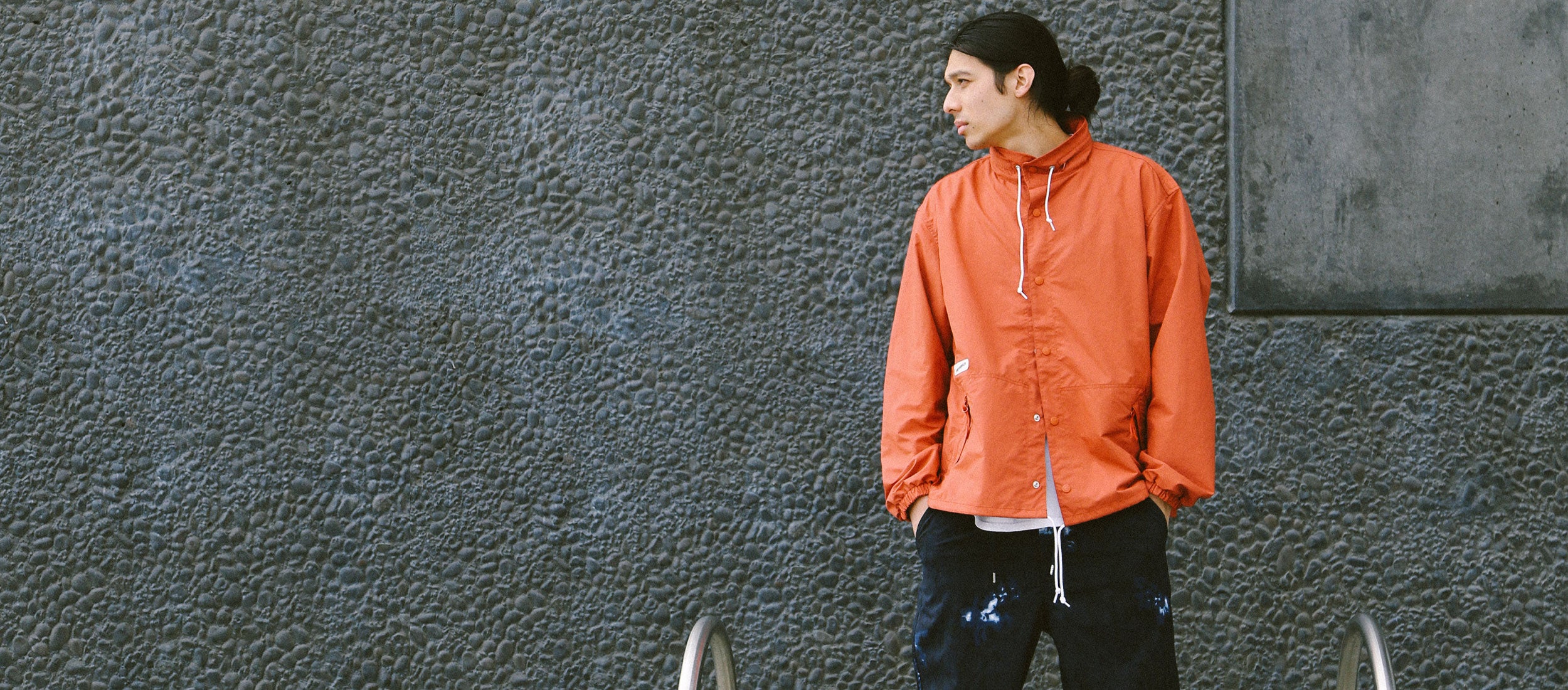 Man in vibrant orange Japanese outdoor jacket and black tie-dye pants, urban texture backdrop, minimalist streetwear style.