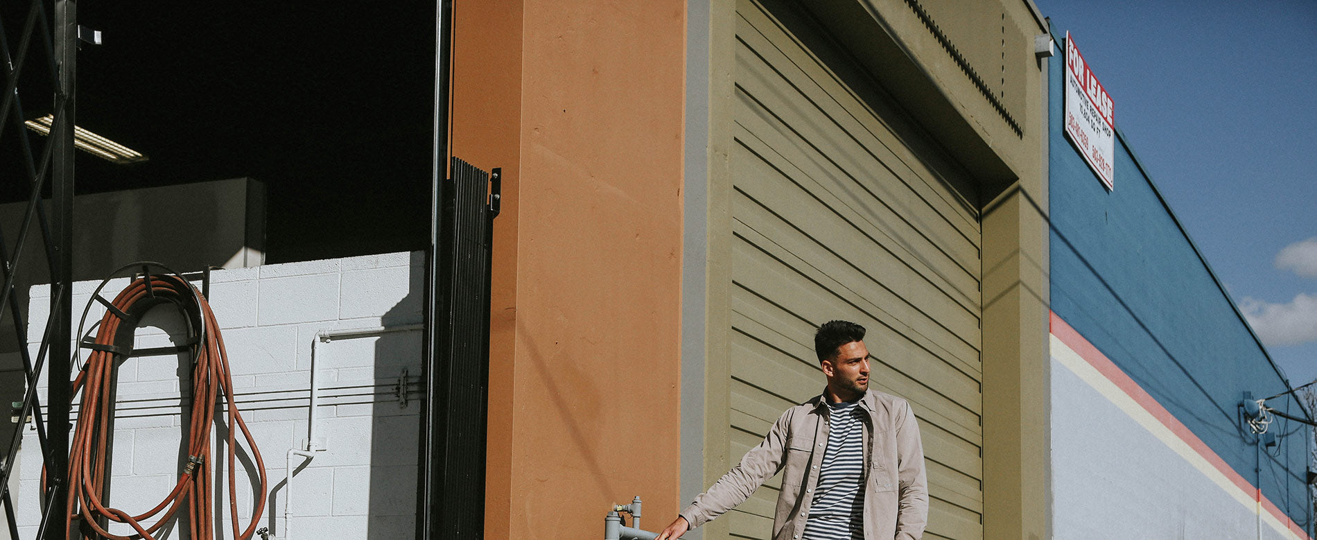 Man in beige jacket and striped tee stands by industrial garage, urban backdrop, Norse Projects minimalist menswear.