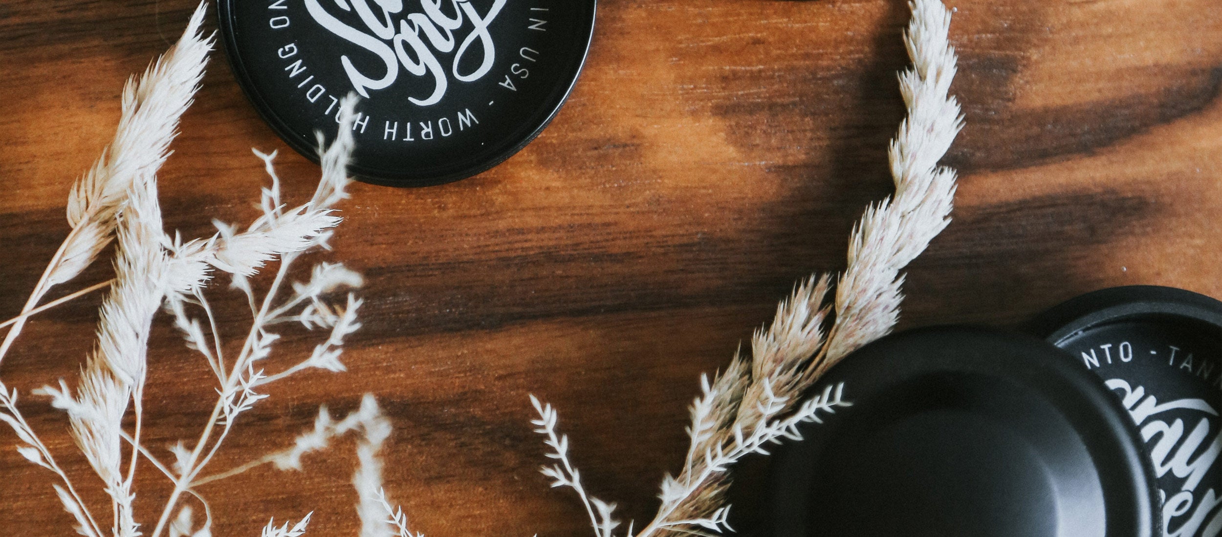 Eco-chic black tins with white script on warm wood, accented by dried grasses—sustainable, minimalist, nature-inspired essentials.