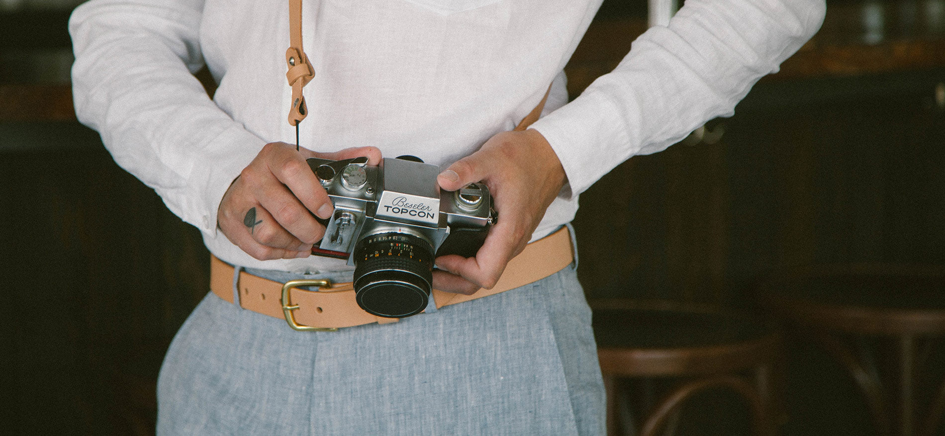 Vintage camera with leather strap, worn by person in white shirt and blue linen pants, evoking timeless wedding photography style.