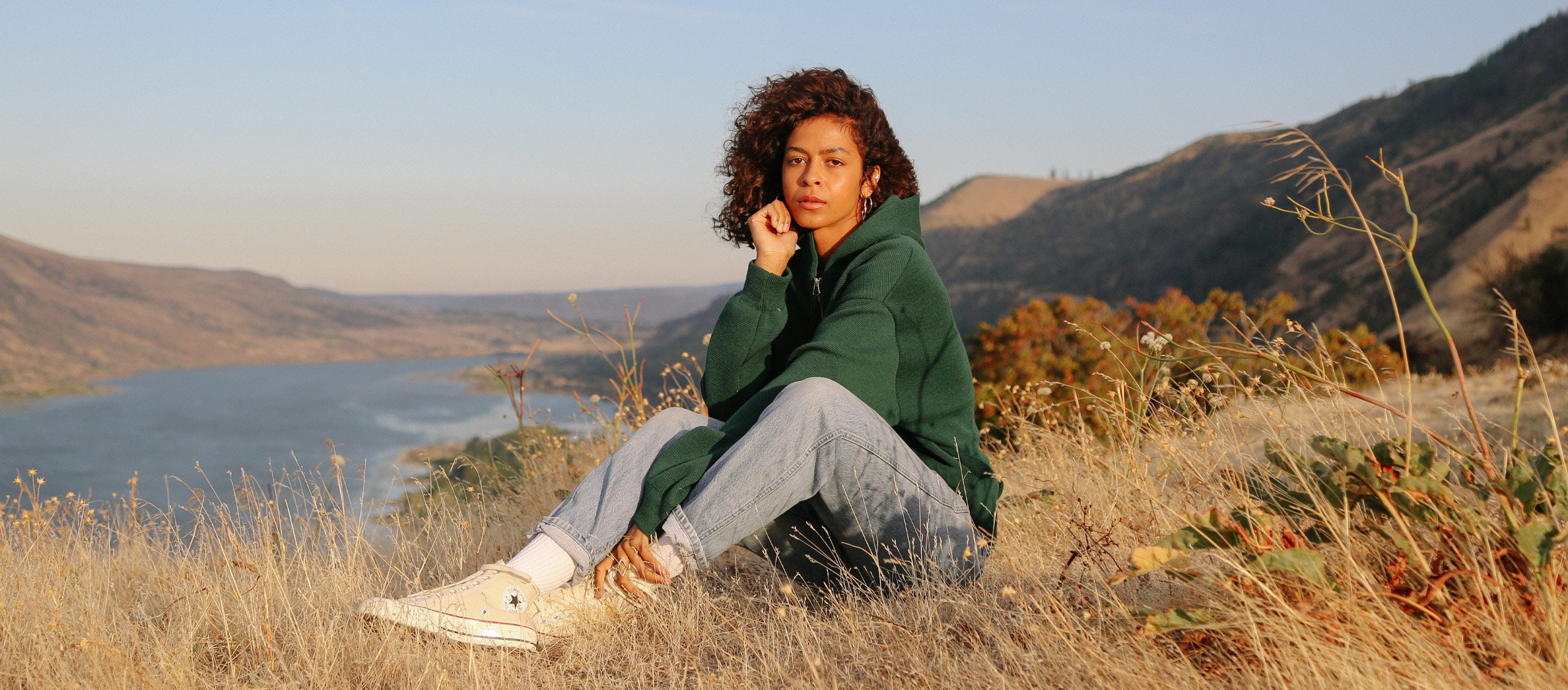 Woman in forest green hoodie and light jeans sits on hillside, overlooking lake and mountains, embodying Arpenteur’s wanderlust-ready, earth-toned outdoor style.