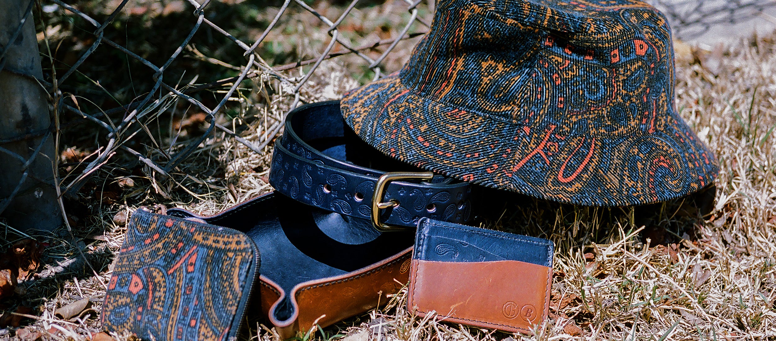 Tanner Goods x Bricks & Wood: rustic leather belt, wallet, and patterned bucket hat in earthy blues, oranges, and browns, styled outdoors for rugged, vintage-inspired menswear.