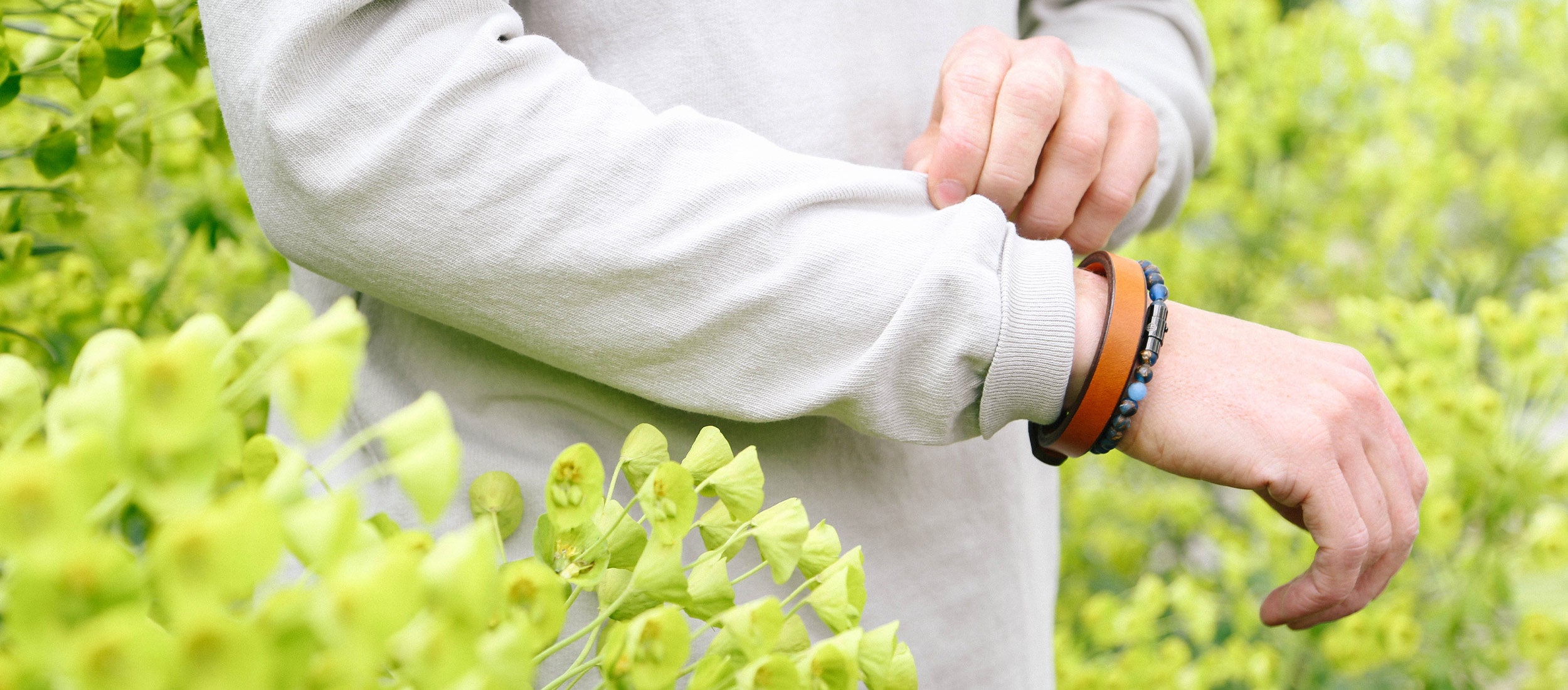 Man’s wrist adorned with leather and beaded bracelets, set against vibrant green foliage, showcasing natural, earthy jewelry.