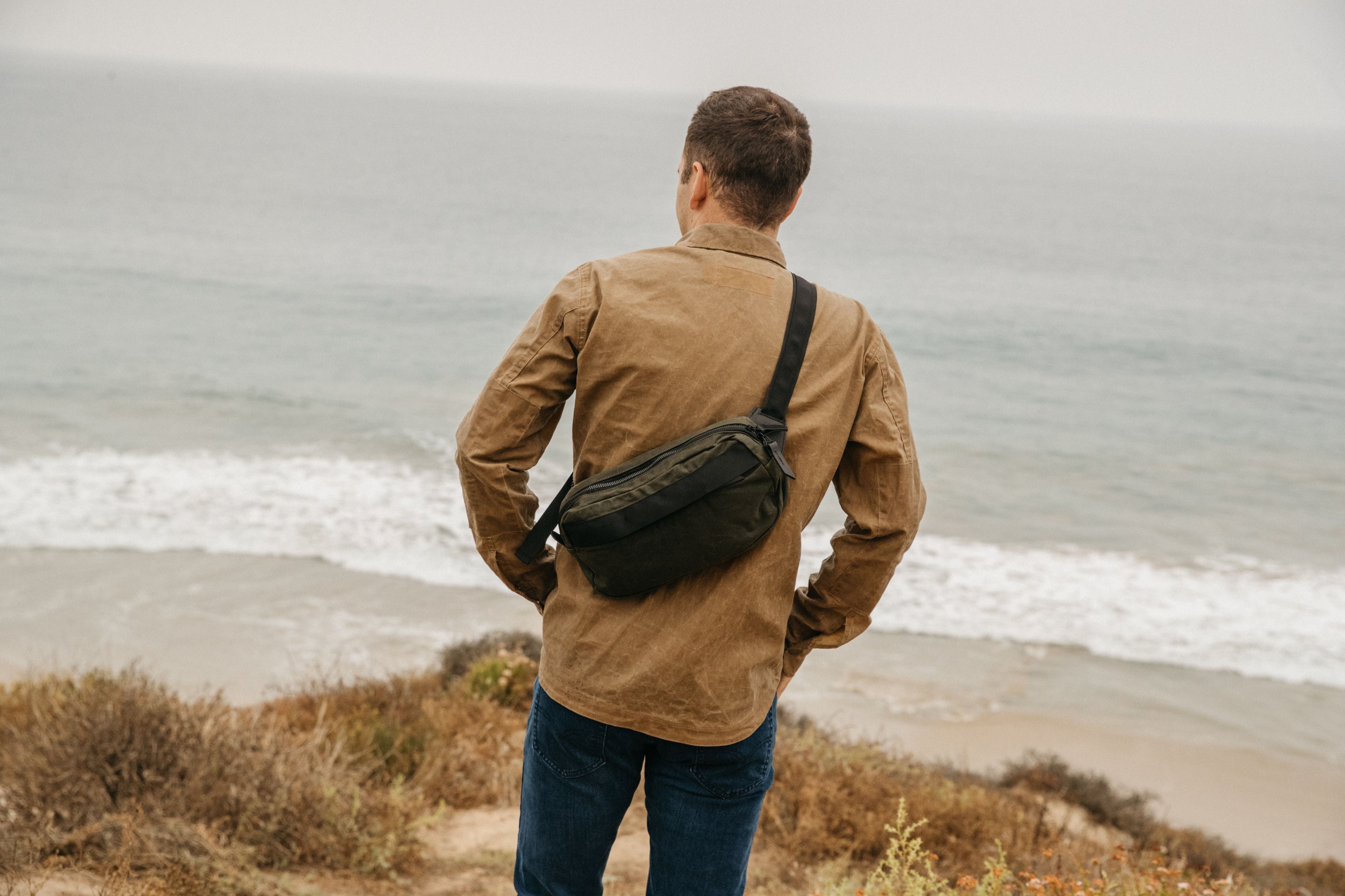 Man wearing olive green waist bag by ocean, rugged outdoor style, earthy tones, adventure-ready.