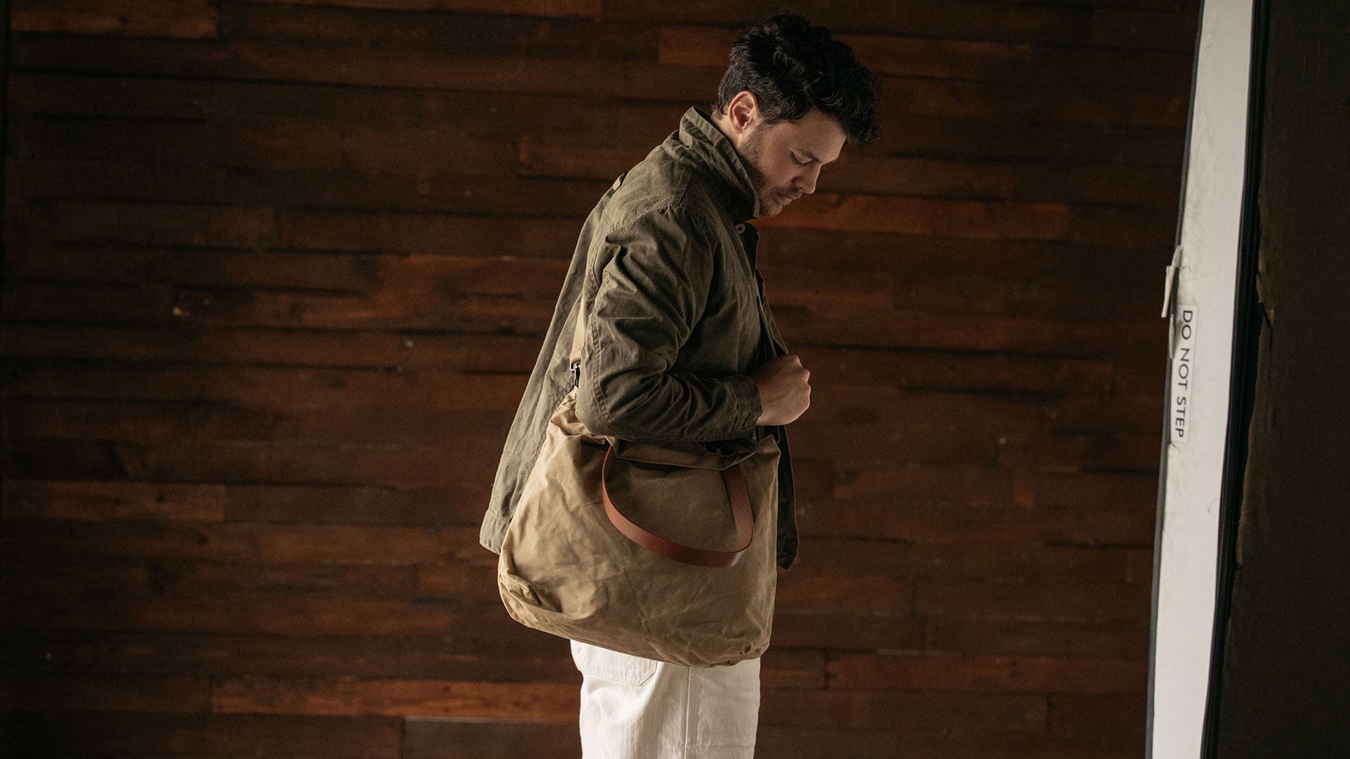 Man in olive jacket carries tan canvas tote with leather strap against rustic wood wall, showcasing durable, minimalist travel essentials.