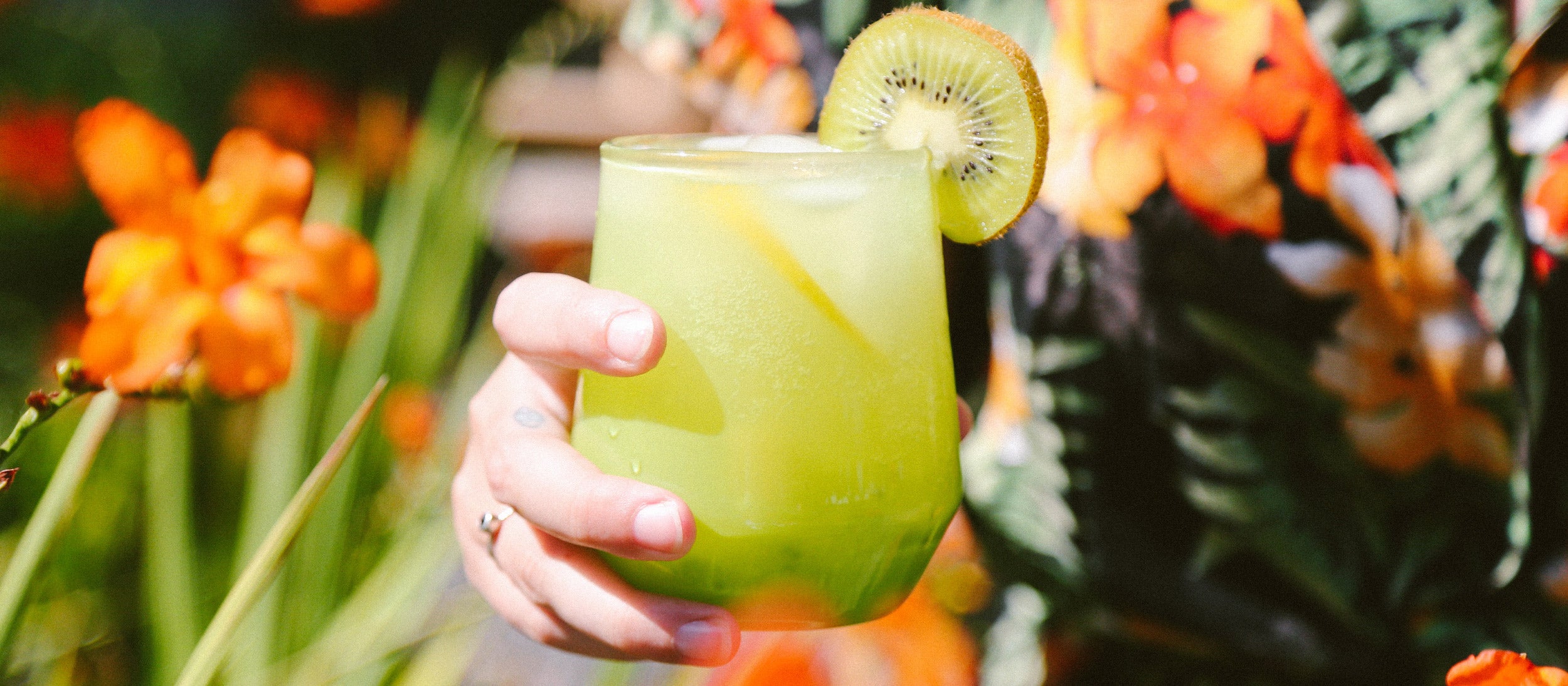 Hand holding vibrant green cocktail in textured glass with kiwi garnish, tropical flowers, and lush foliage—Summer Glassware collection.