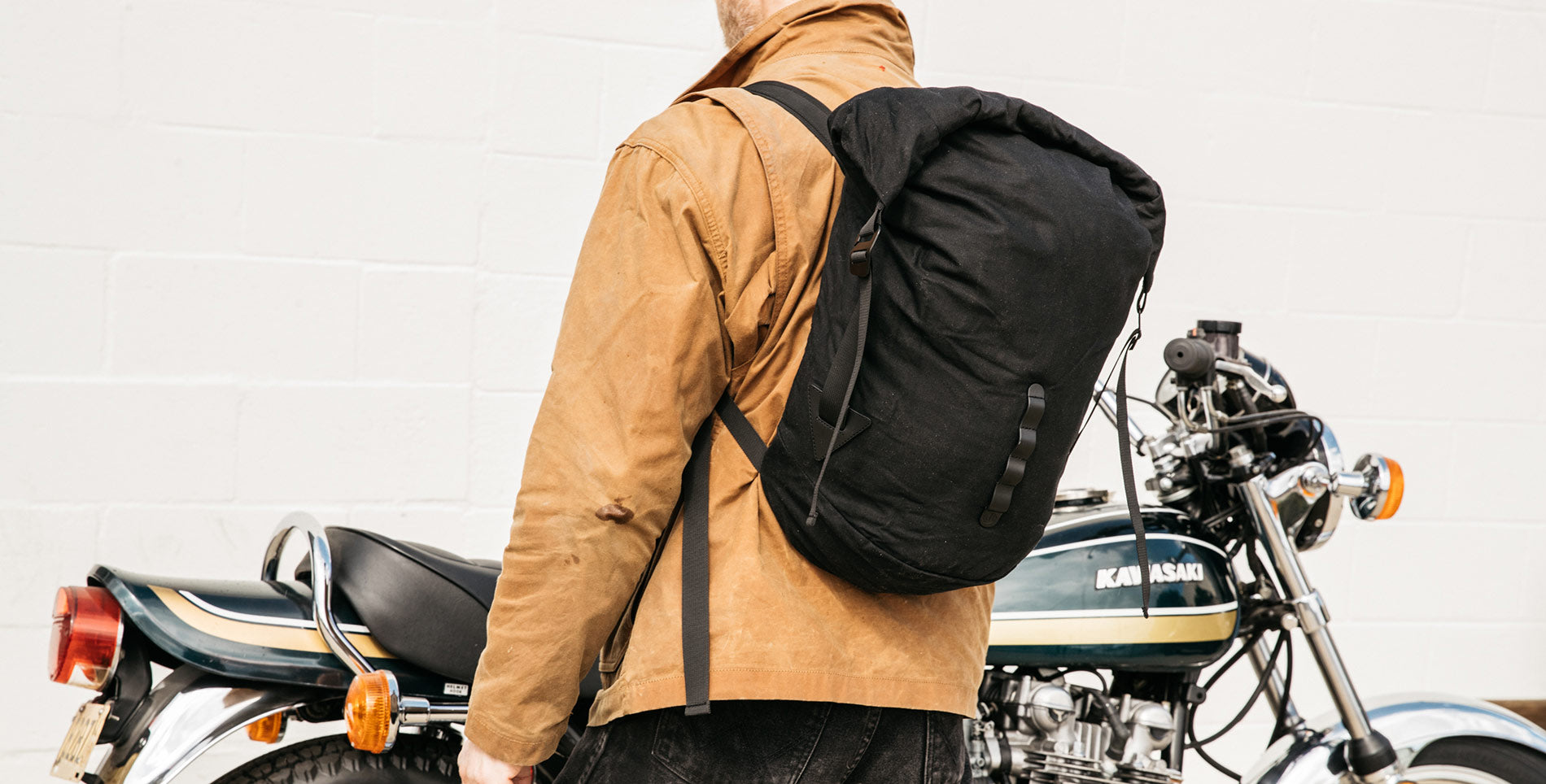 Man in tan jacket with black backpack stands beside vintage Kawasaki motorcycle, rugged urban style.