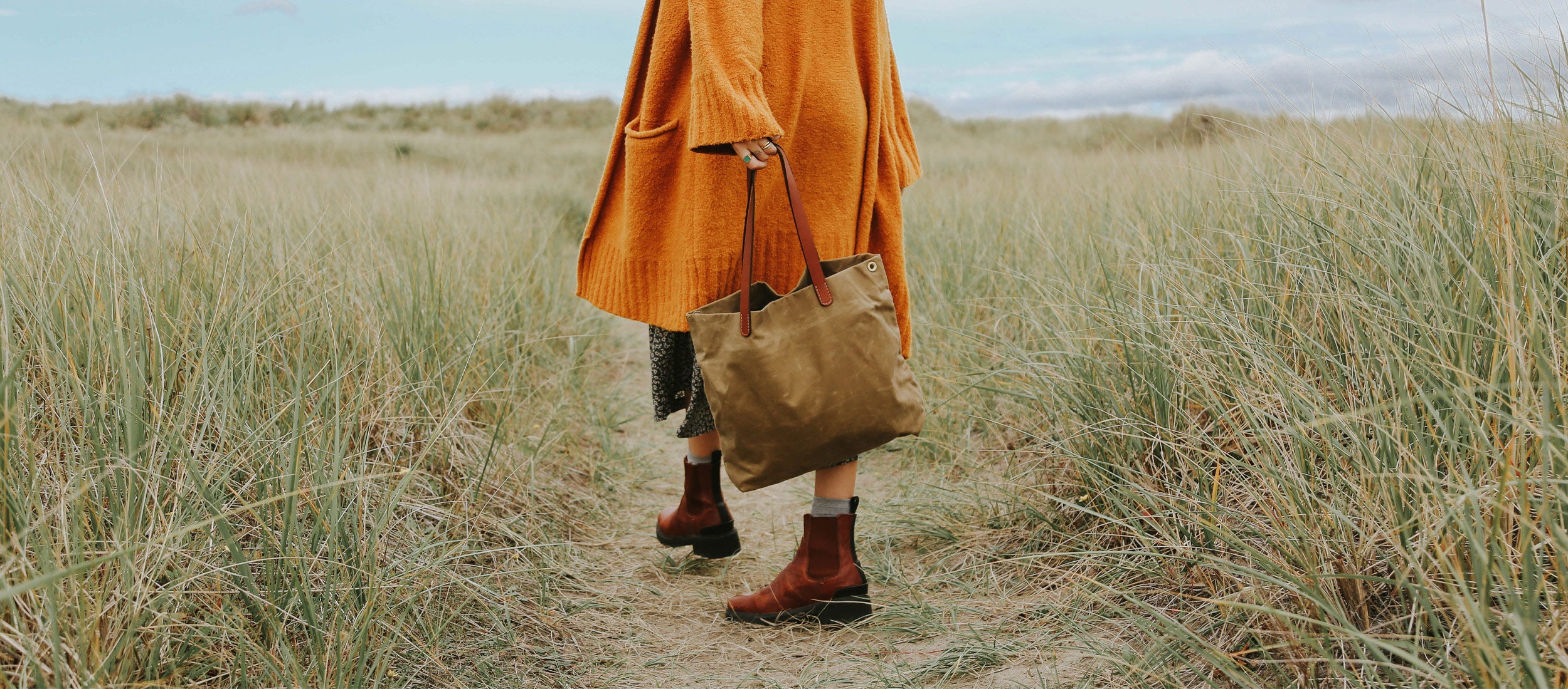 Woman in orange coat carries tan canvas tote through coastal grasses, earthy tones, rustic style, sustainable everyday bags.