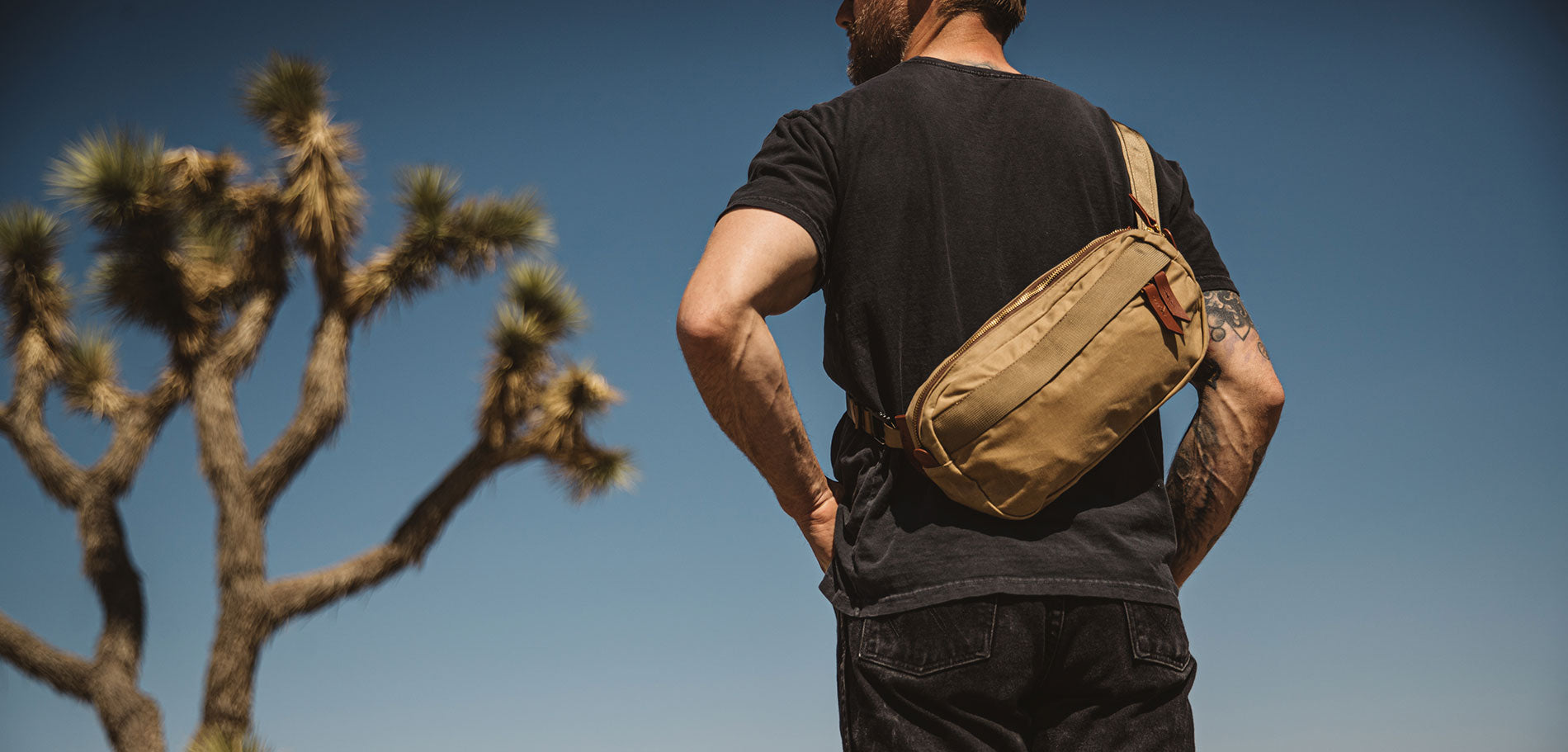 Man in desert landscape wearing tan utility waist bag, black tee, and jeans under clear blue sky.