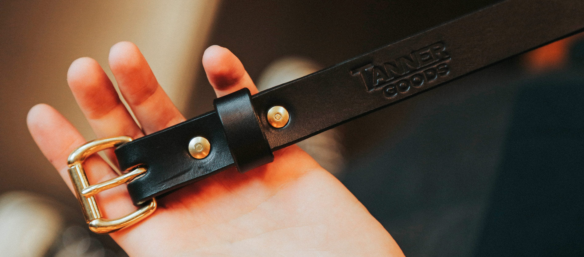 Black leather skinny belt with gold hardware, embossed Tanner Goods logo, held in hand — sleek, minimalist style.