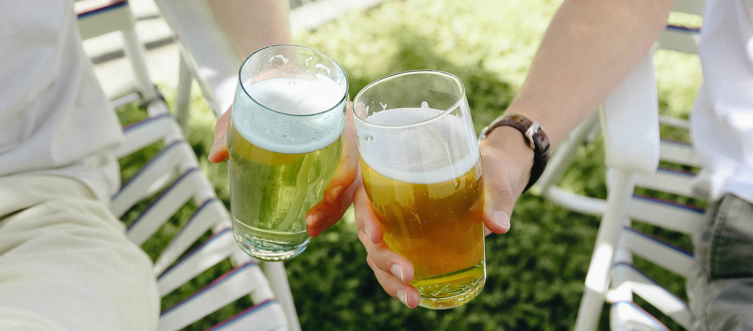 Two hands clinking beer glasses in a sunny backyard, white chairs and green grass, relaxed summer vibe.