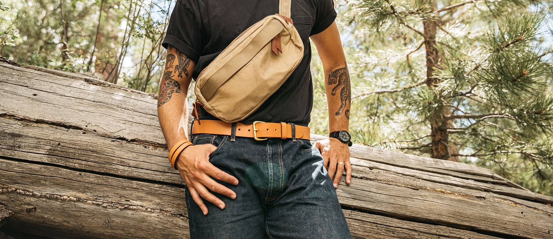Man in forest wearing tan leather belt, matching bracelet, and canvas bag—rustic, rugged style.