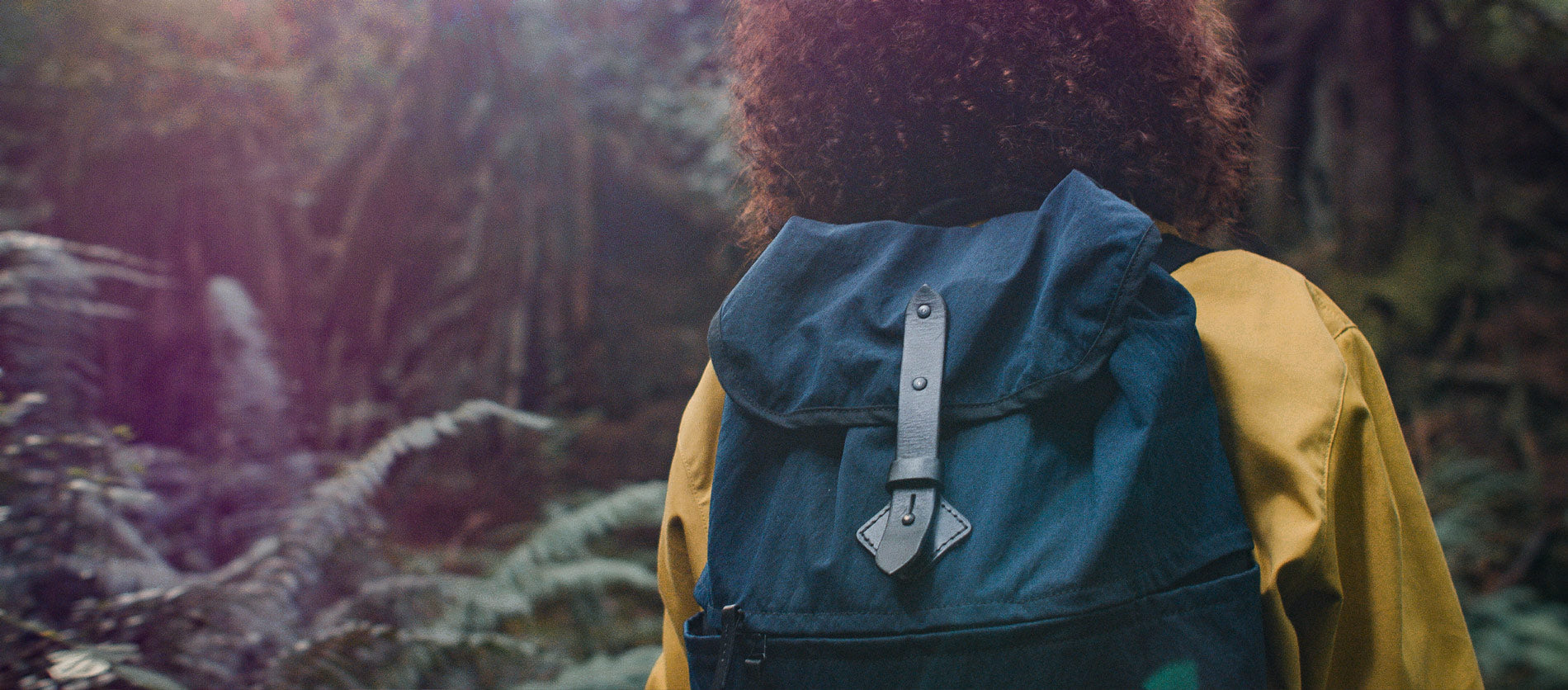 Person wearing navy Voyager daypack with leather buckle, hiking through misty forest, earthy tones, adventure-ready style.