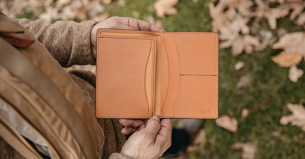 Hands holding open tan leather full-size wallet with card slots, outdoors on mossy ground.