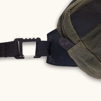Juniper green waxed canvas crossbody bag with a black webbing strap, featuring a unique black metal buckle and leather accent, in a cropped view.