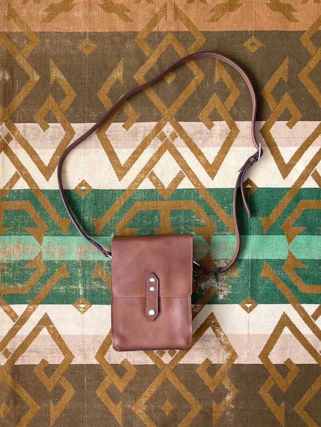 Brown leather crossbody bag with a flap closure and adjustable strap, laid flat on a geometric patterned blanket.