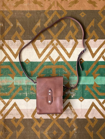 Brown leather crossbody bag with a flap closure and adjustable strap, laid flat on a geometric patterned blanket.