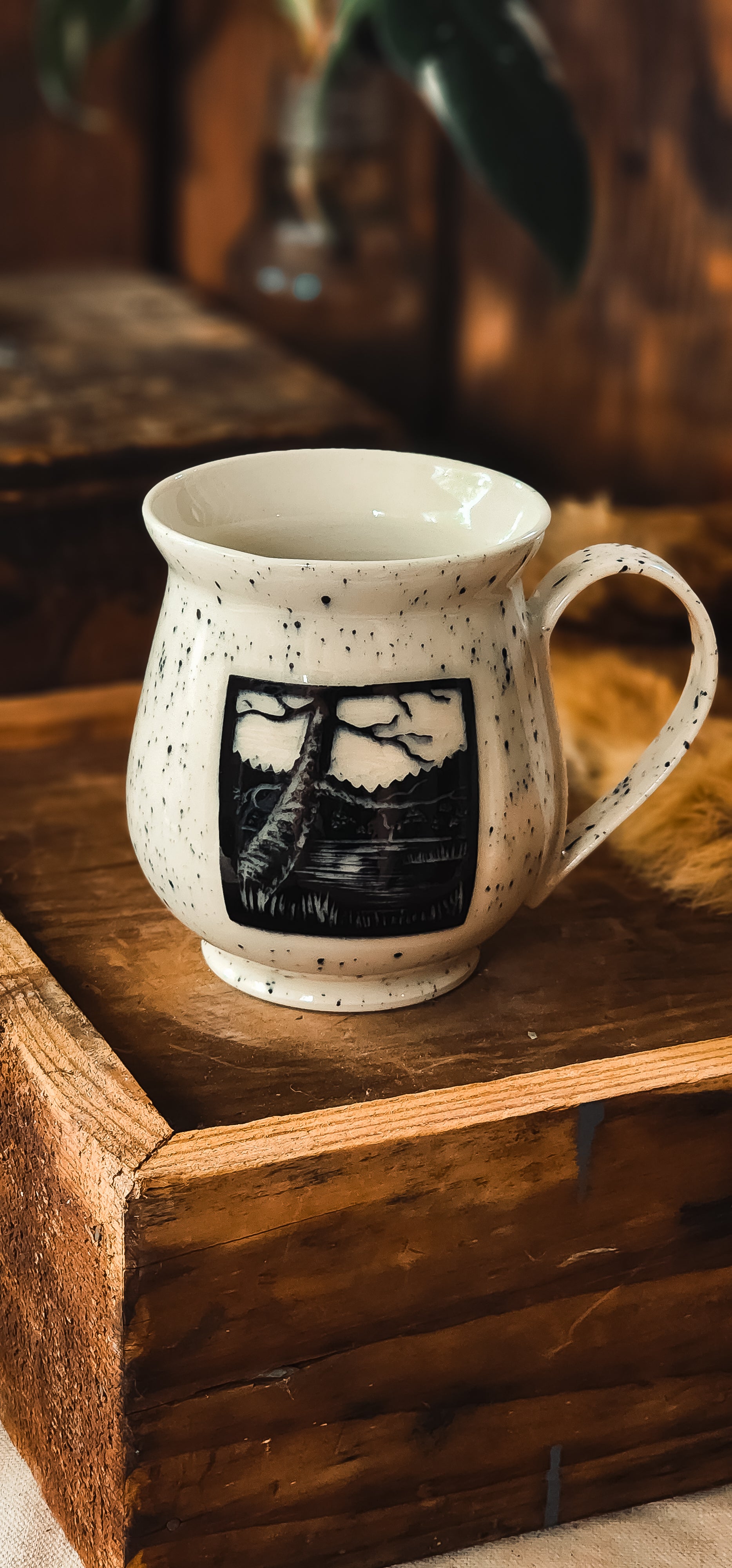 White ceramic mug with black speckles, featuring a black and white lake and tree design, resting on a wooden crate.