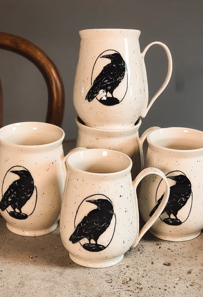 Cream speckled ceramic Raven Mug with a black raven design in a circle, displayed with multiple stacked mugs.