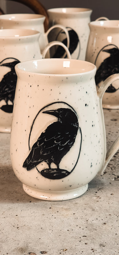 Off-white speckled mug featuring a prominent black raven design inside a circular outline, with blurred mugs in the background.